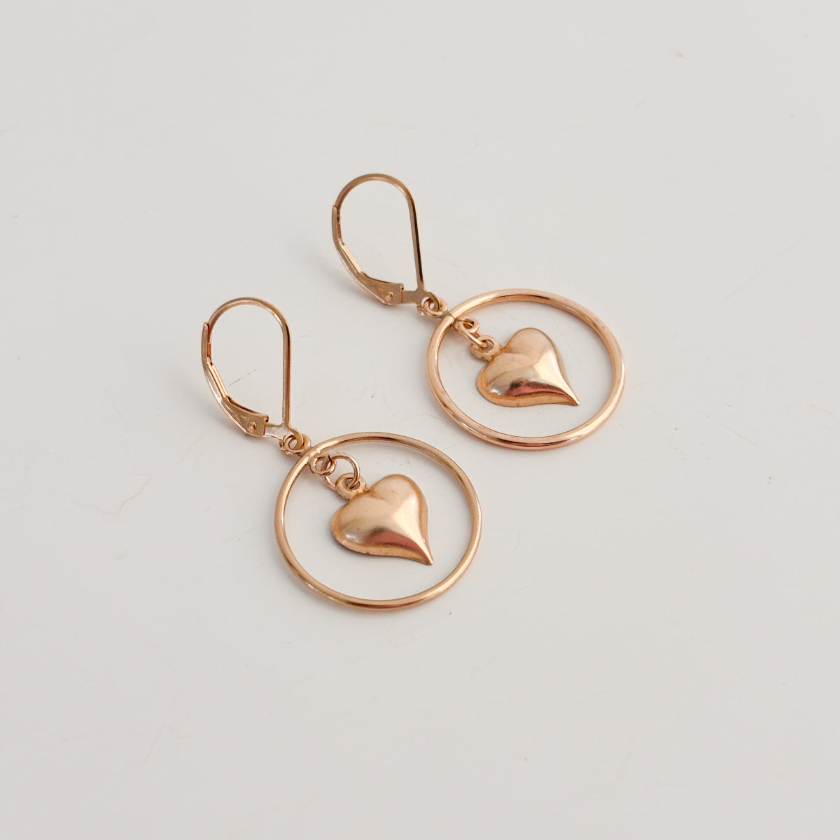 a pair of lever back earrings in 14k gold filled metal.  The earrings feature an open circle hoop frame and a dangling puffy heart charm within the circle. 