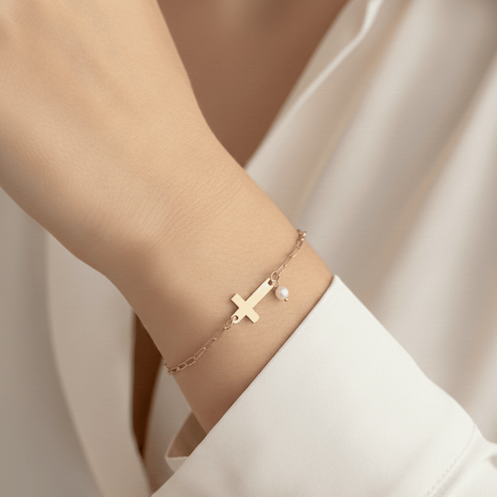 Gold bracelet with a cross and pearl on a wrist against a neutral background