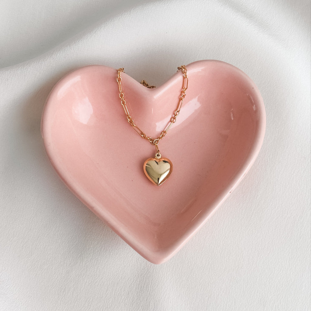 a 14k gold-filled long and short chain necklace that features a large puffy heart pendant shown in a pink heart-shaped ceramic dish.