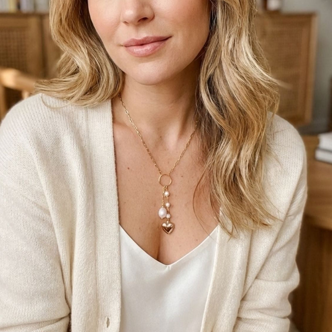 Woman wearing a gold paperclip necklace with duo pendants featuring a gold puffed heart charm and pearlsin a cozy indoor setting