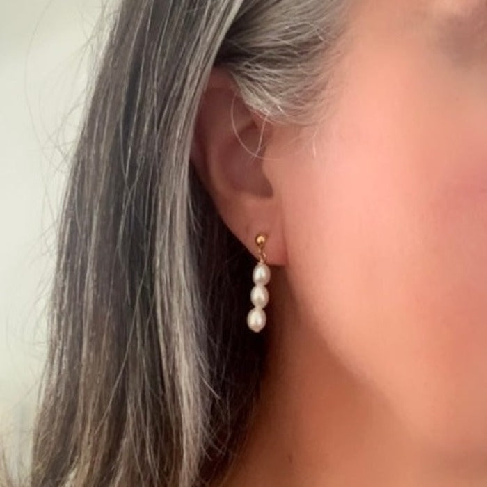 a single pearl earring shown on ear.  The earring features a drop of three small freshwater pearls that dangles from a gold ball stud earring. 