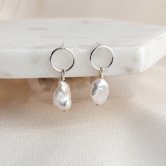 Open circle post earrings featuring baroque keshi pearl drops  on a marble stand with a neutral background