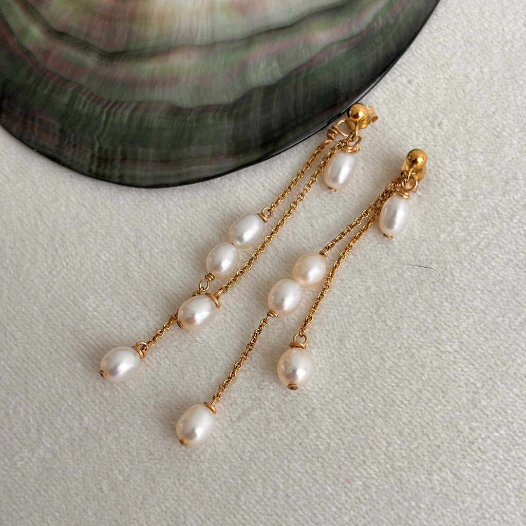 Pearl earrings with gold chains on a light gray surface shown next to a shell