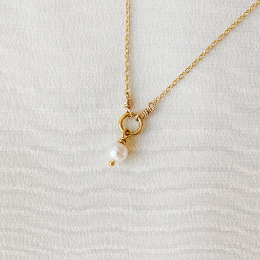 Gold necklace with a central open ring from which dangles a single petite pearl pendant on a light gray background