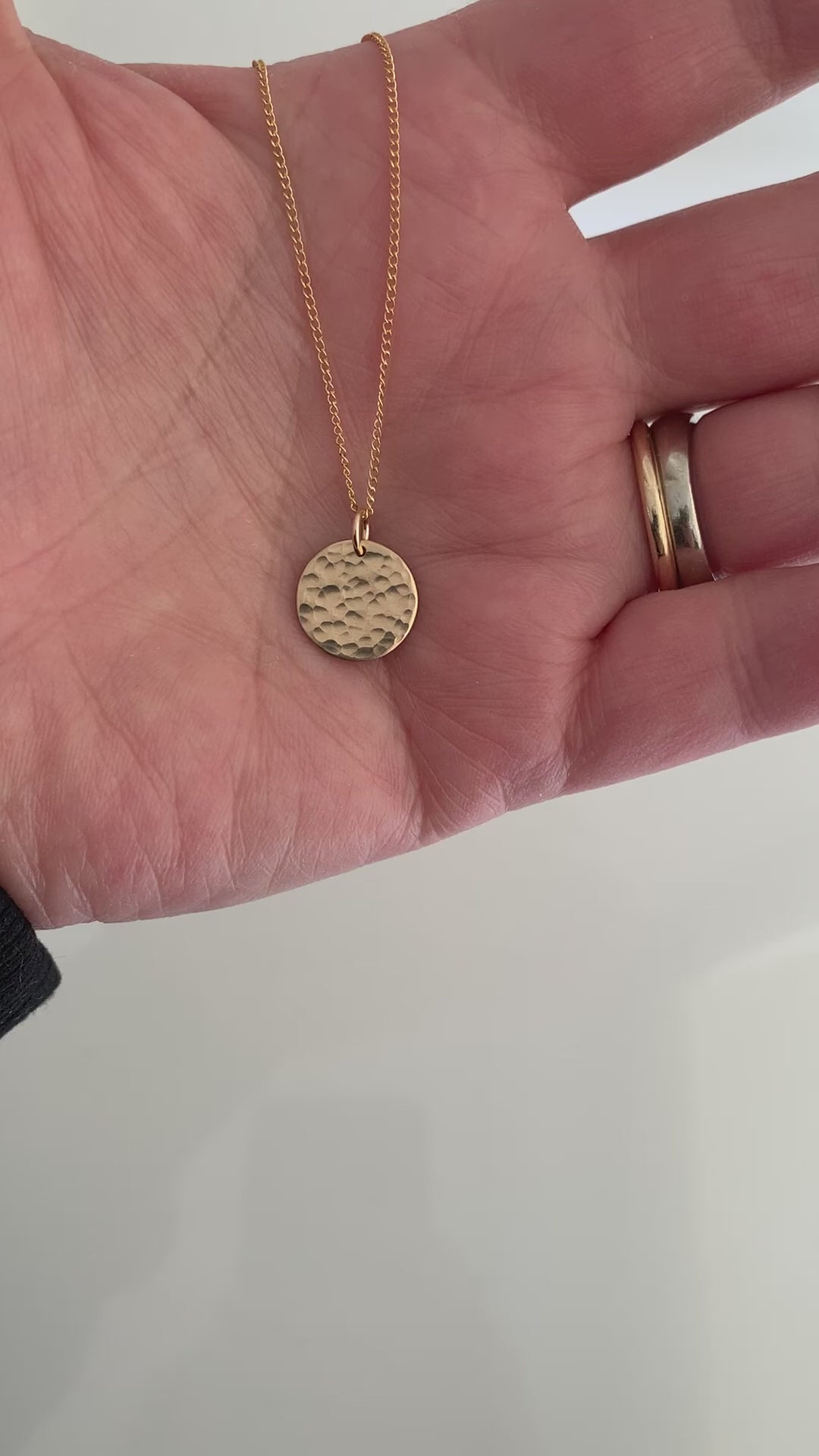 A short video clip of a 14k gold filled hammered disc pendant necklace on a fine curb chain shown held in a hand .  