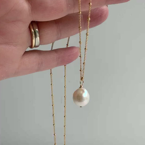A gold pearl necklace shown dangling from a hand. The large white Edison pearl is off round with some texture and high metallic sheen. The pearl has a hand wired loop and is suspended from 14k gold filled beaded chain.