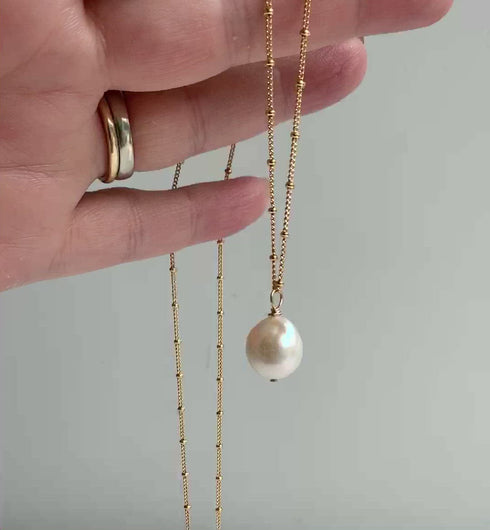 A gold pearl necklace shown dangling from a hand. The large white Edison pearl is off round with some texture and high metallic sheen. The pearl has a hand wired loop and is suspended from 14k gold filled beaded chain.